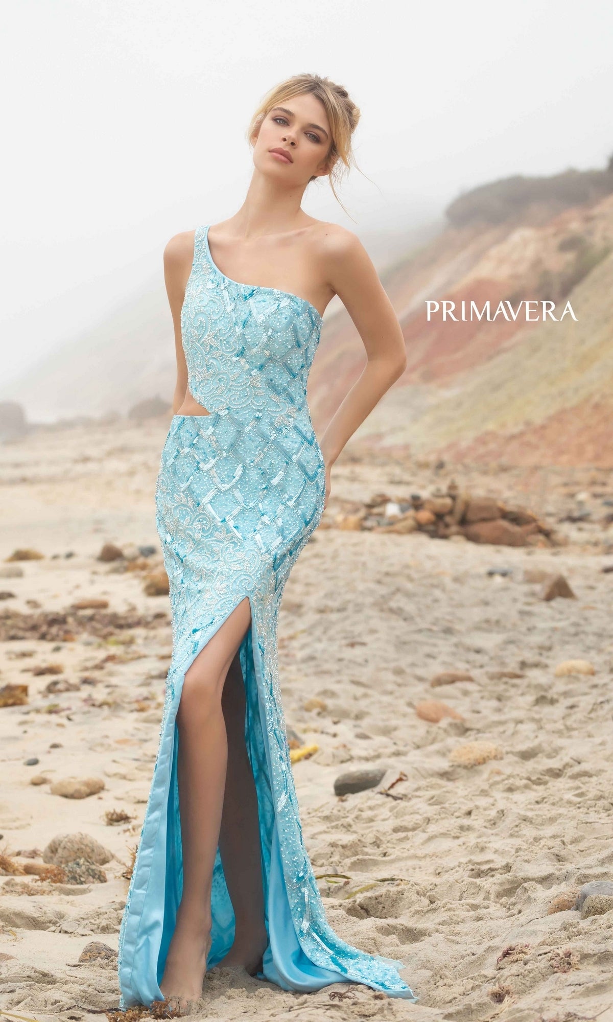 A woman wears the Primavera 3729 One-Shoulder Long Sequin Prom Dress in light blue, featuring a thigh-high slit, as she stands barefoot on a sandy beach with rocky cliffs.